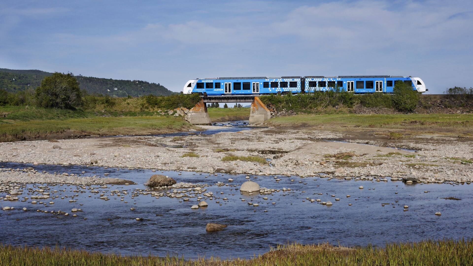 blue and white hydrogen powered train travelling over small bridge over stream 