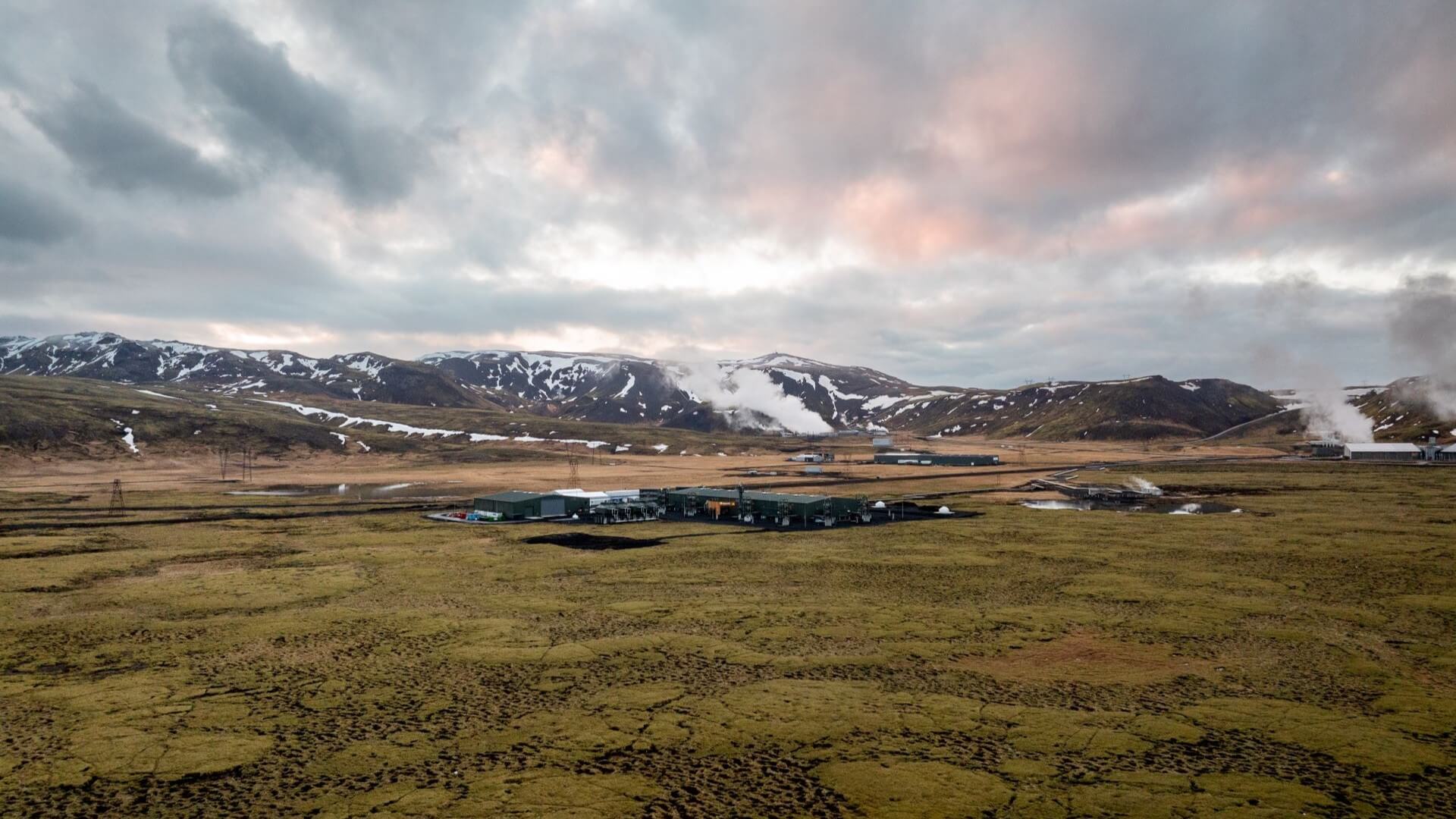 view of industrial plant set in green scrubby grass and snow covered mountains behind, with geothermal steam venting in background