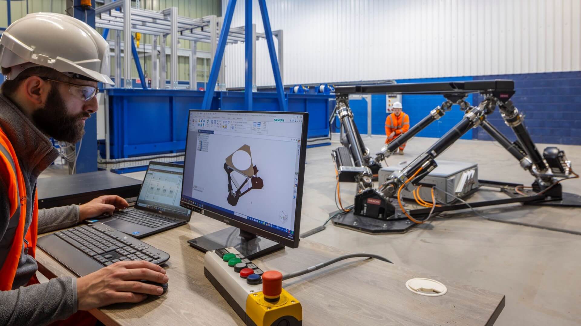 man in high viz jacket and hard hat sat in front of laptop showing CGI image of 3D triangular hexapod structure, replicating the real-life structure on the workshop floor in front of him