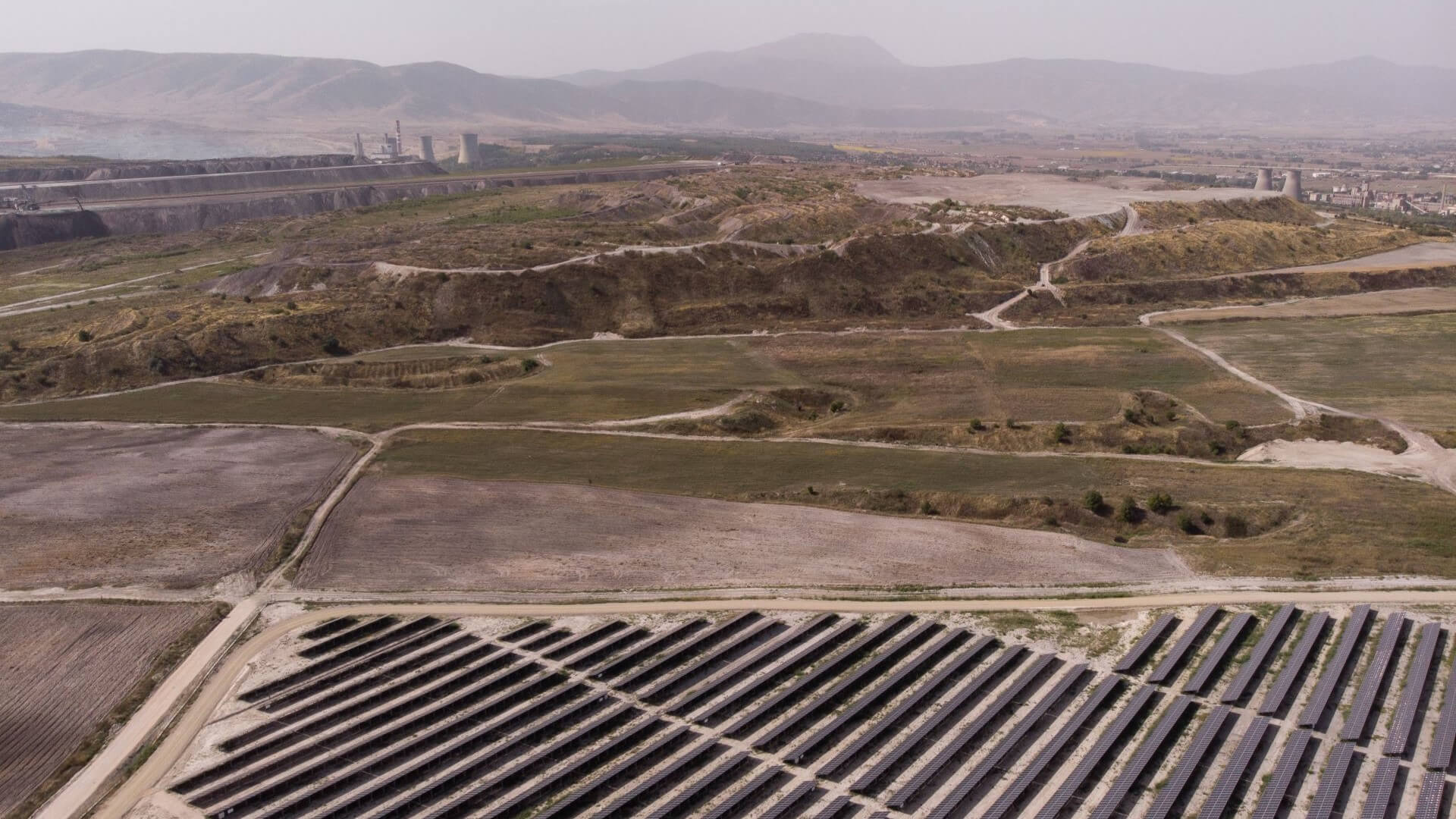 rows of solar panels in foreground with rough landscape and industrial buildings in background