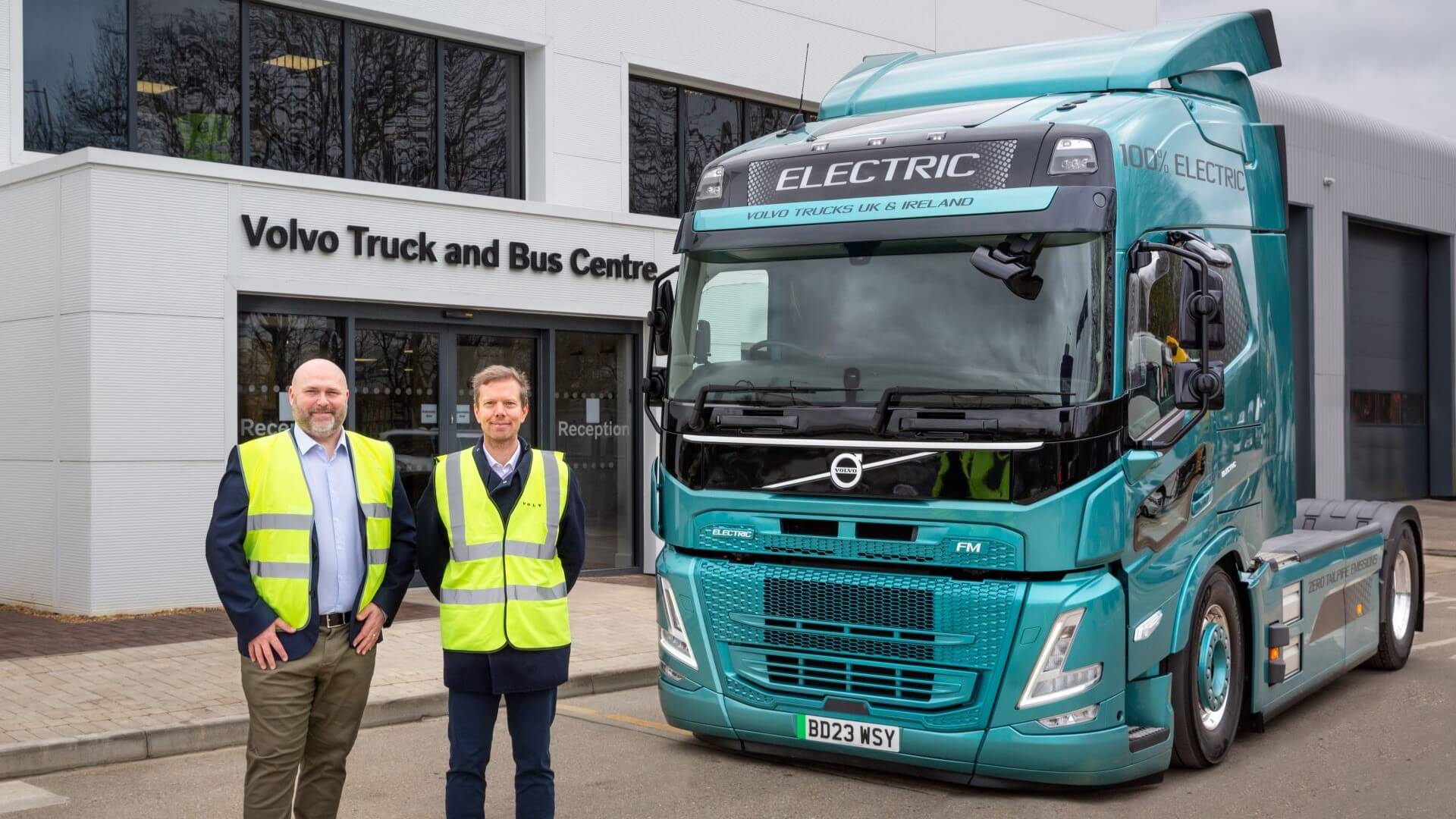 two men in high vis jackets standing next to large truck next to front of building