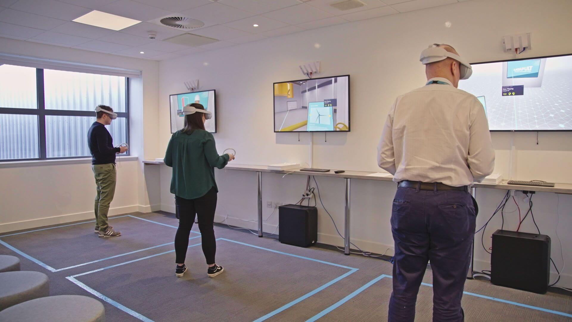 three people wearing virtual reality headsets, standing in a line across the room, in front of three screens projecting images