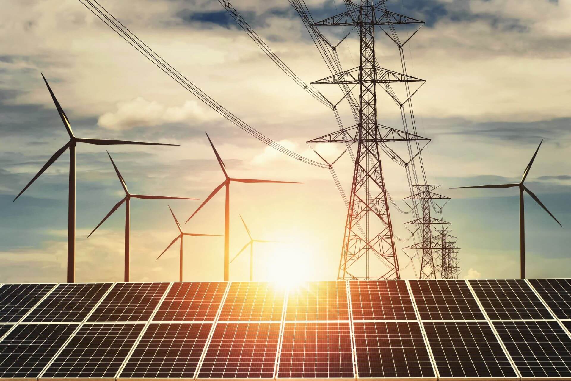 rows of solar panels in foreground with wind turbines behind to left and electricity pylons to right behind set against rising sun and skyline